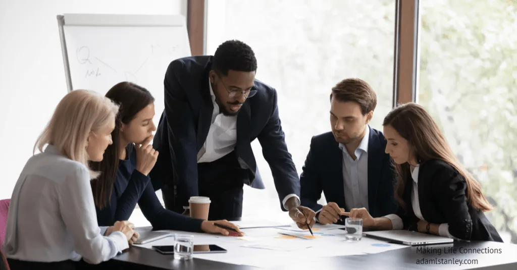 Black professional man standing over a table explaining something to a mixed group of other professionals, all wearing dark suits and lightly colored shirts.
