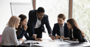 Black professional man standing over a table explaining something to a mixed group of other professionals, all wearing dark suits and lightly colored shirts.
